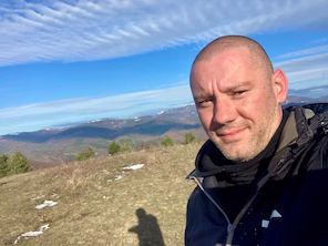 Ivaylo Milkov on a grassy mountain summit with the mountain range behind him.
