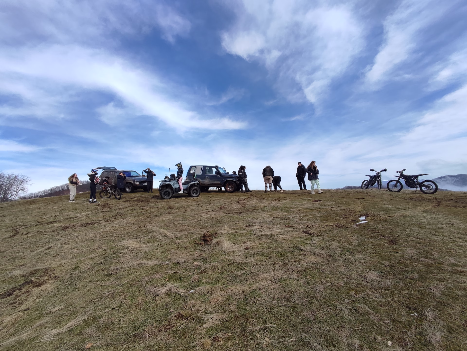 A wide group of off-road enthusiasts gathered on a grassy hilltop under a dramatic blue sky with wispy white clouds, with 4x4 SUVs, an ATV, electric dirt bikes, and people spread across the ridge.