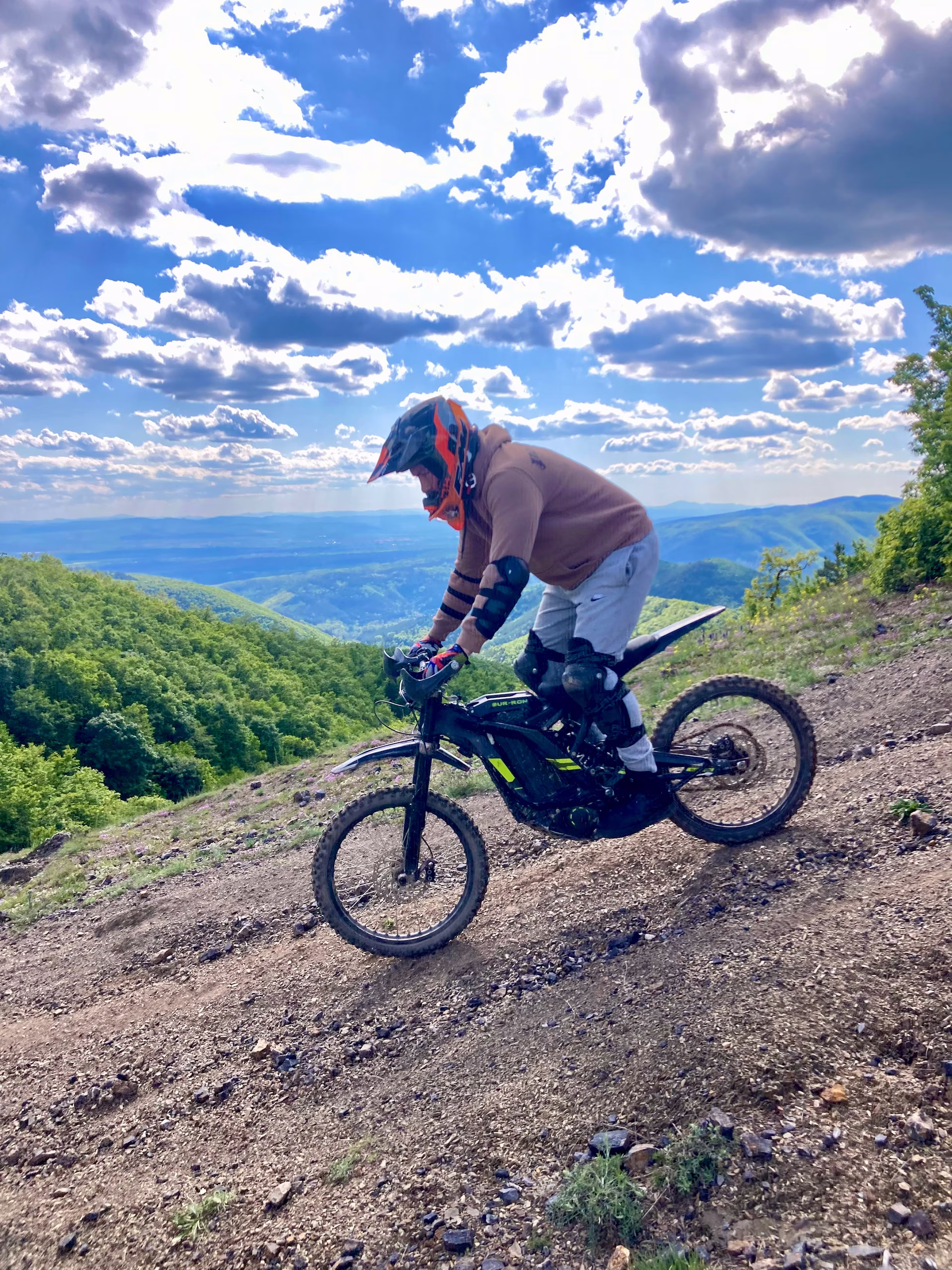A rider in a helmet, hoodie, and full elbow and knee pads descending a steep, gravelly mountain trail on a black electric dirt bike, with vibrant green forests and a cloudy blue sky in the background.