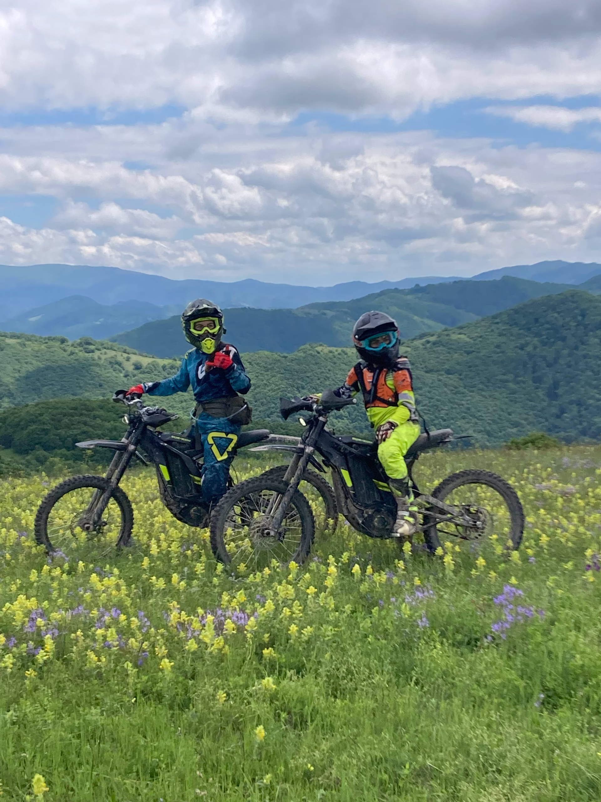 Two young off-road riders in full gear and helmets sitting on black electric dirt bikes in a field of wild yellow flowers, with lush green mountain ridges in the background.