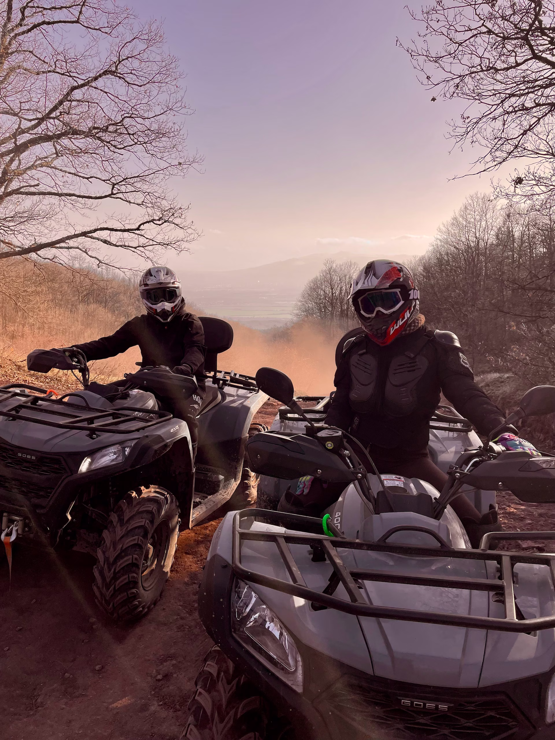Two off-road riders wearing helmets and protective suits riding grey ATV quad bikes along a forest dirt trail during sunset.
