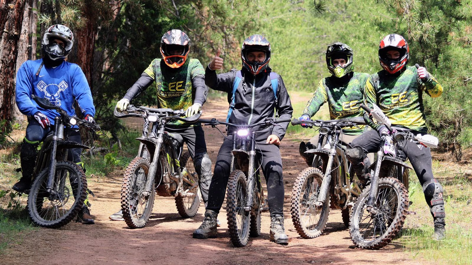 Five off-road riders in full gear and helmets posing together with their electric dirt bikes on a sunny forest dirt trail, with two riders giving a thumbs-up.