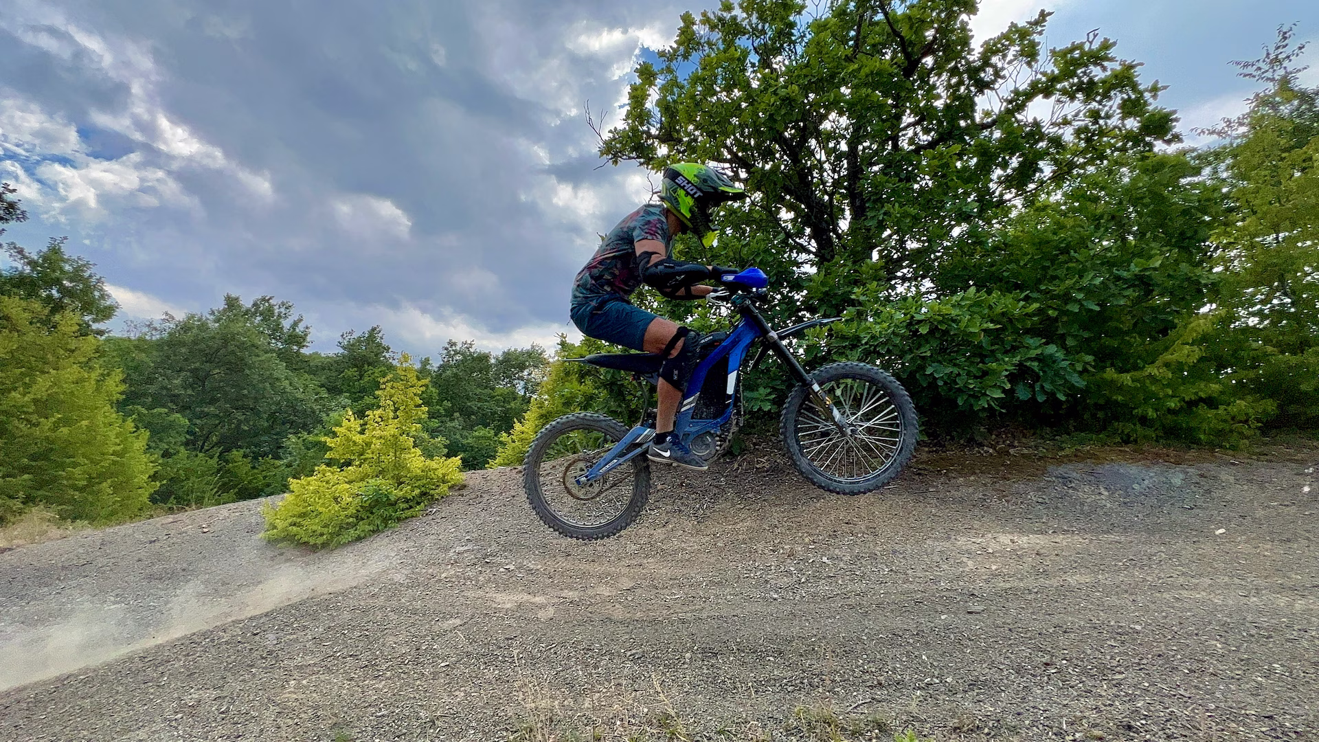 A rider wearing a lime green helmet, elbow pads, and knee pads riding a blue electric dirt bike up a dirt incline surrounded by lush green trees under a cloudy sky.